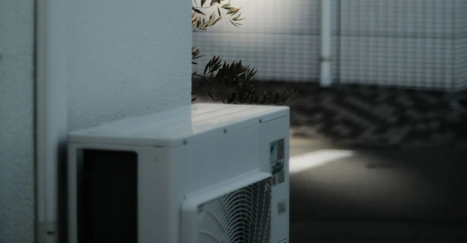 An outdoor air conditioning unit at the corner of a white wall in dappled shade